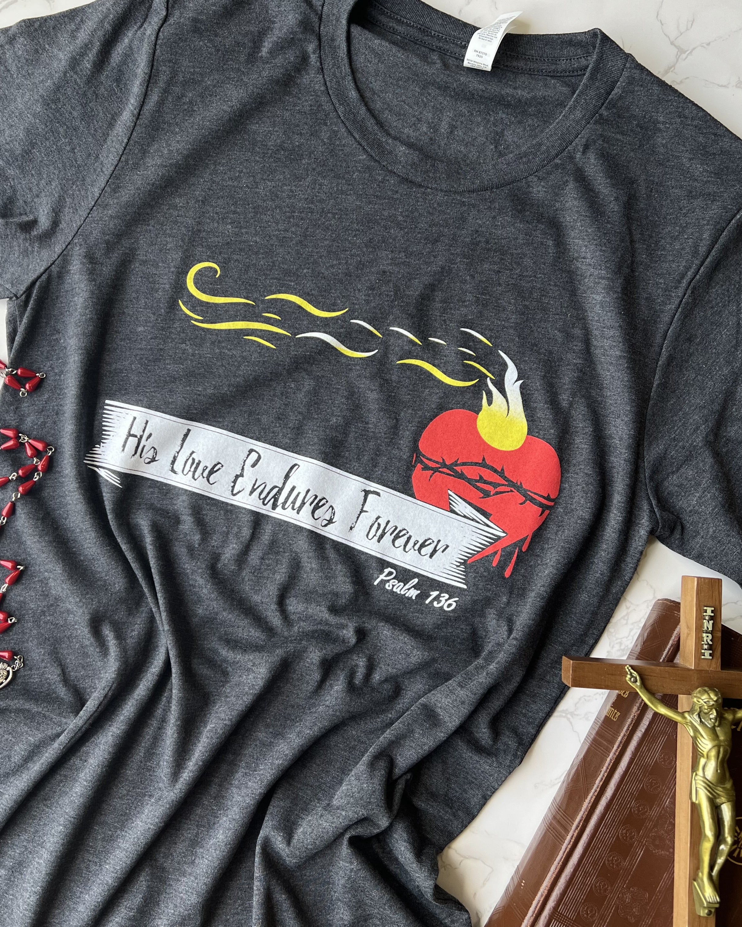 A navy-colored T-shirt with a graphic print of a heart and flames, along with the text 'HIS LOVE ENDURES FOREVER' and the year '187'.