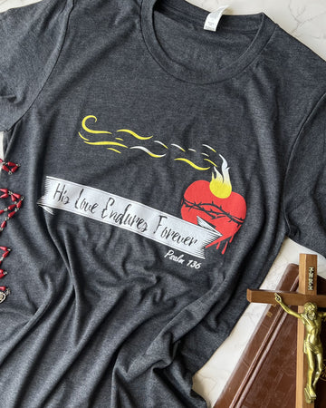 A navy-colored T-shirt with a graphic print of a heart and flames, along with the text 'HIS LOVE ENDURES FOREVER' and the year '187'.