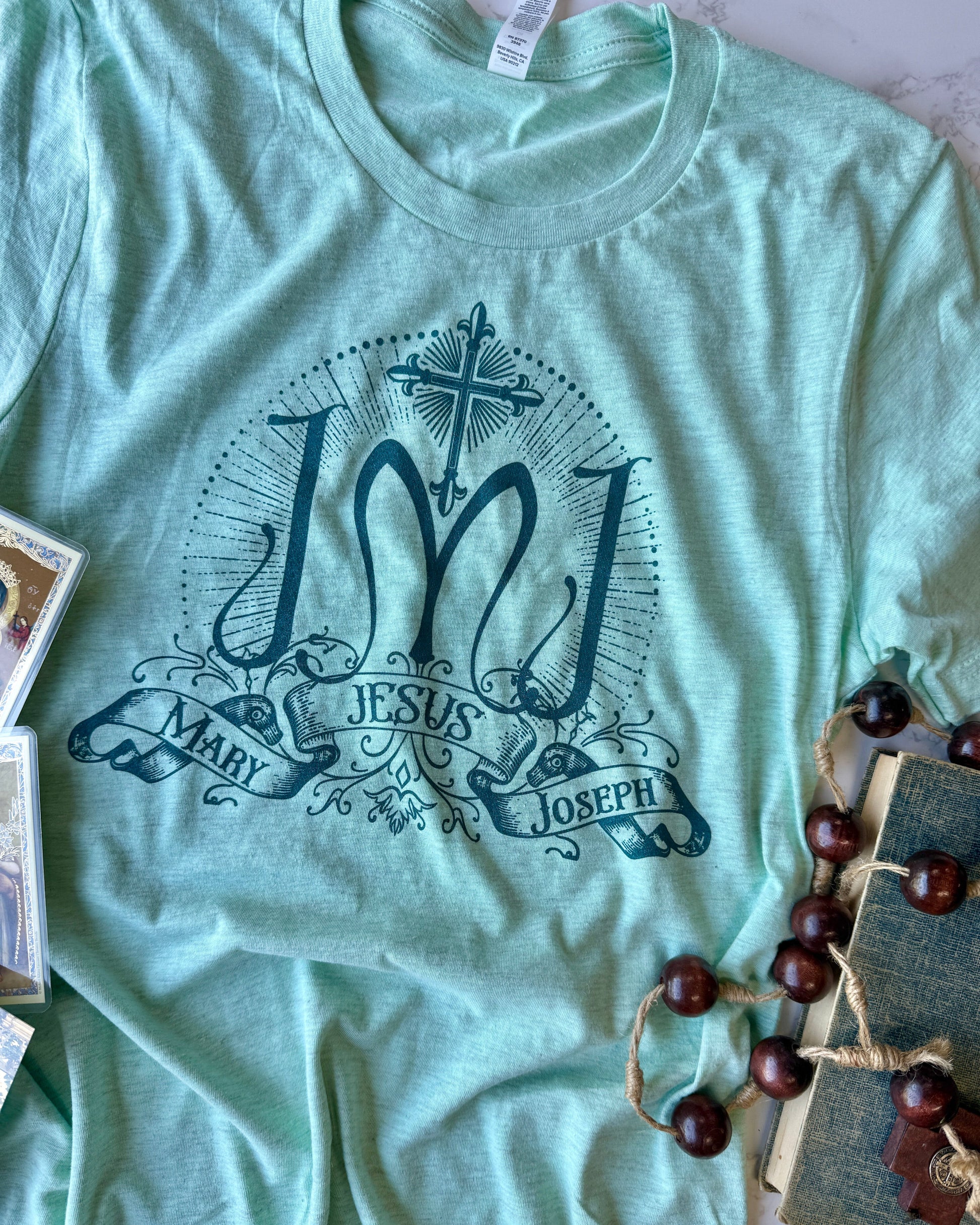 Light green t-shirt with 'Jesus Mary Joseph' design on a surface with decorative items.