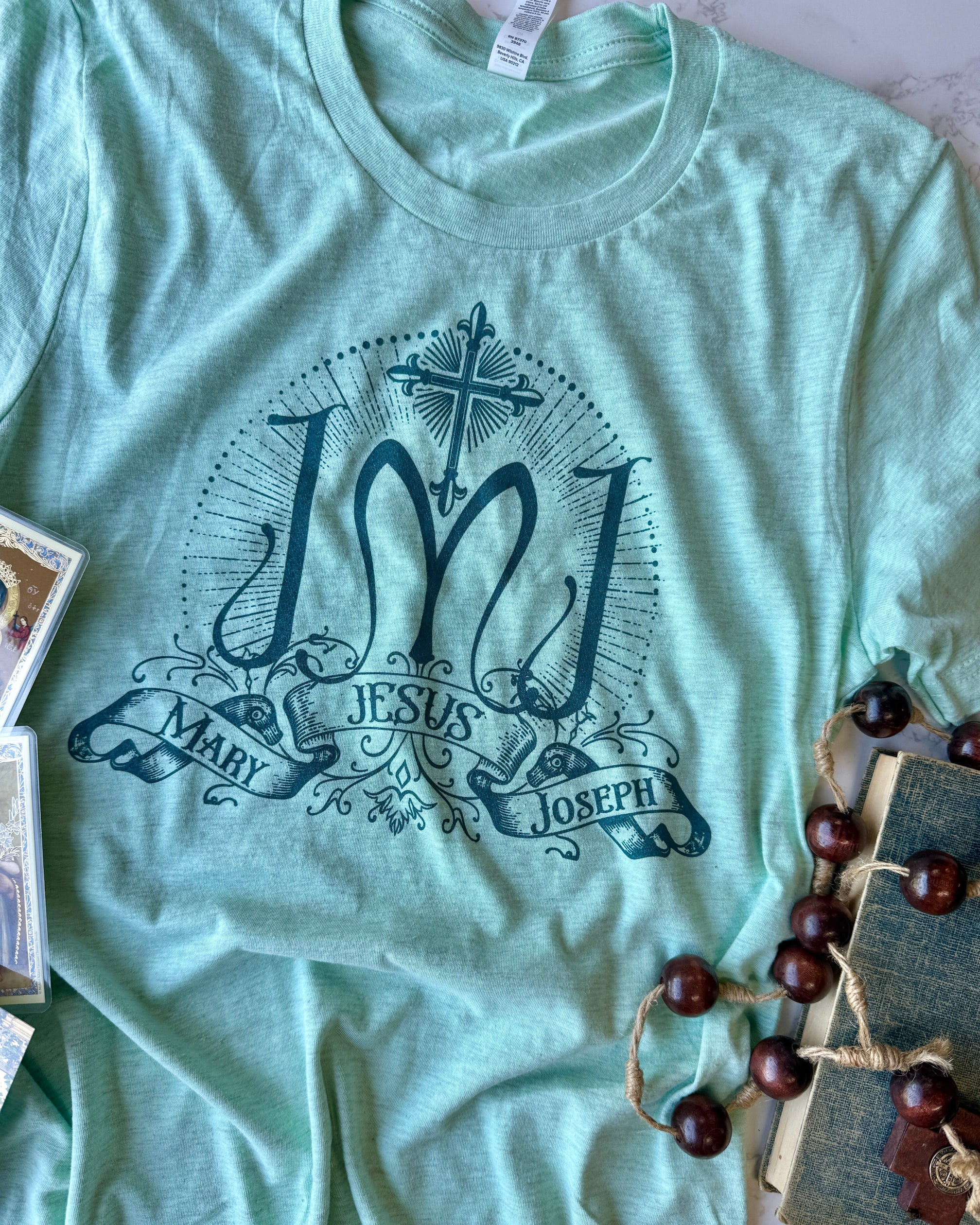 Light green t-shirt with 'Jesus Mary Joseph' design on a surface with decorative items.